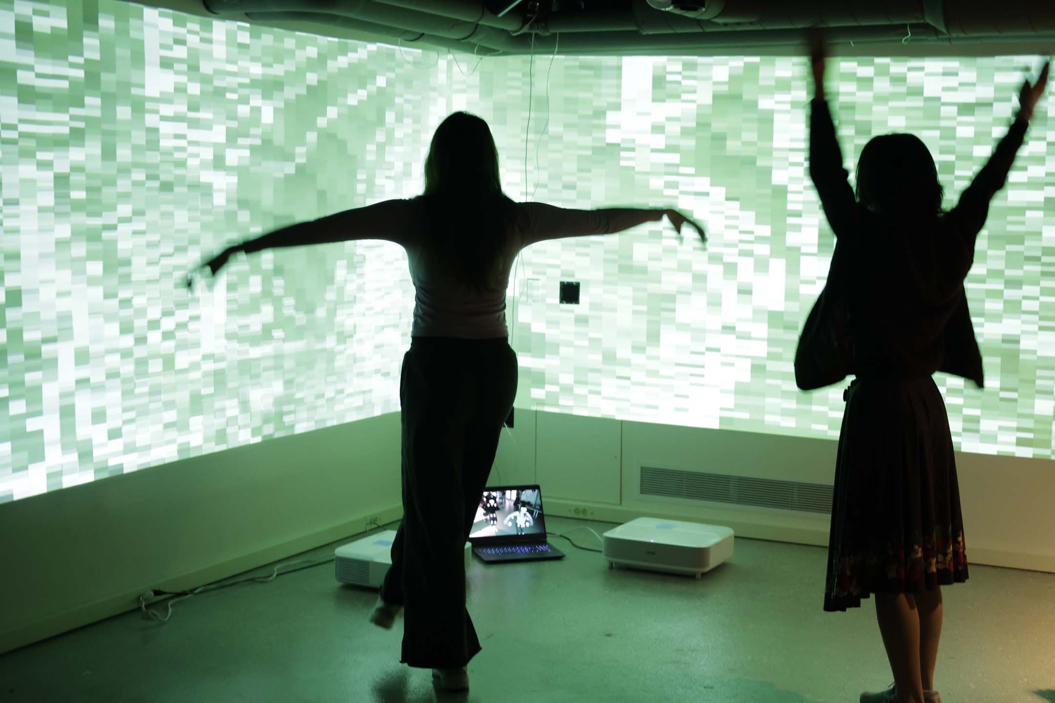 Two people standing in front of a large projection, one is making a T-pose, and the other has their arms in the air. The projection shows a distorted abstract visual that is green and heavily pixelated.