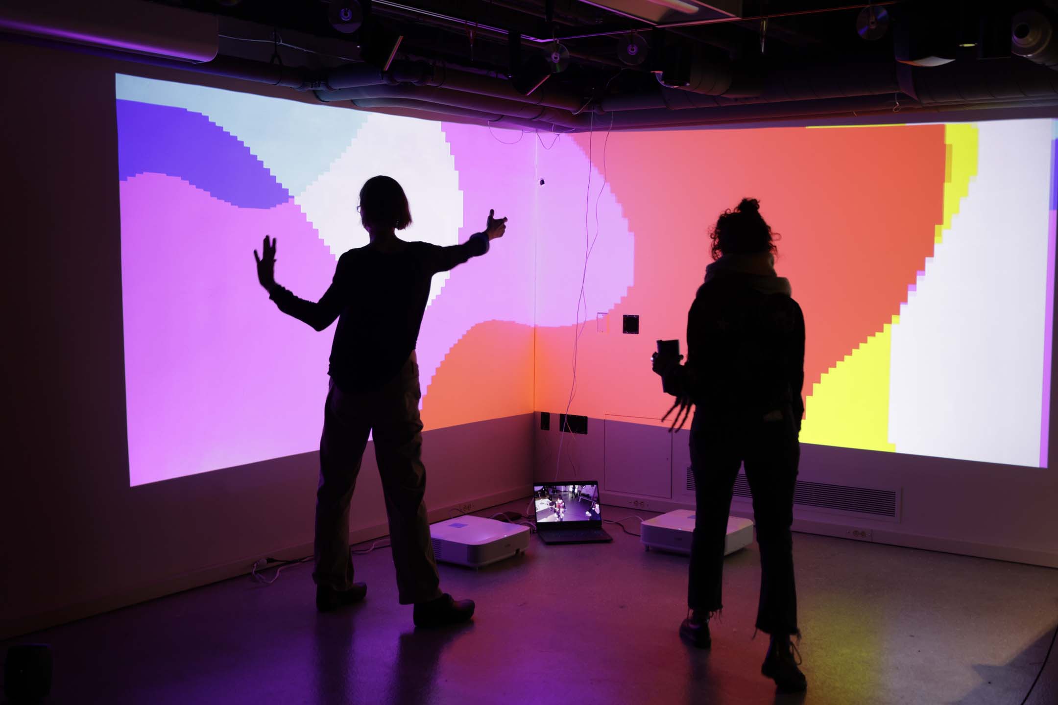 Two people silhouetted against a pixelated RGB background projection, making dynamic poses.