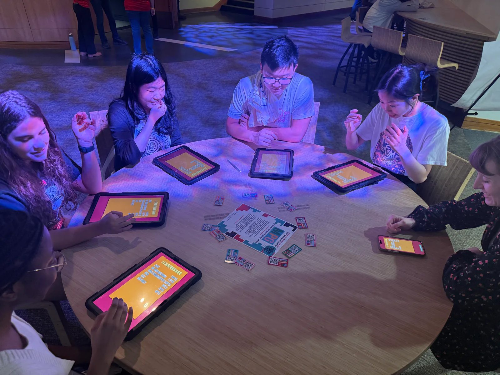 Six people sitting around a round table at NextNOW Fest, each with a phone or tablet in front of them, playing MPG together.