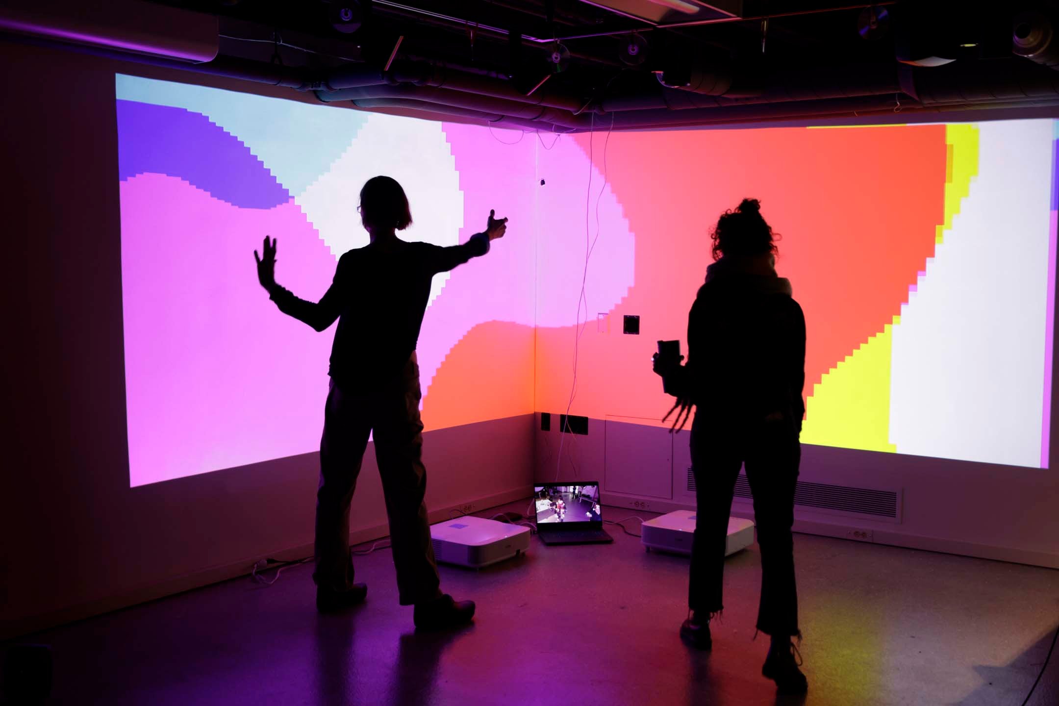 Two people silhouetted against a pixelated RGB background projection, making dynamic poses.