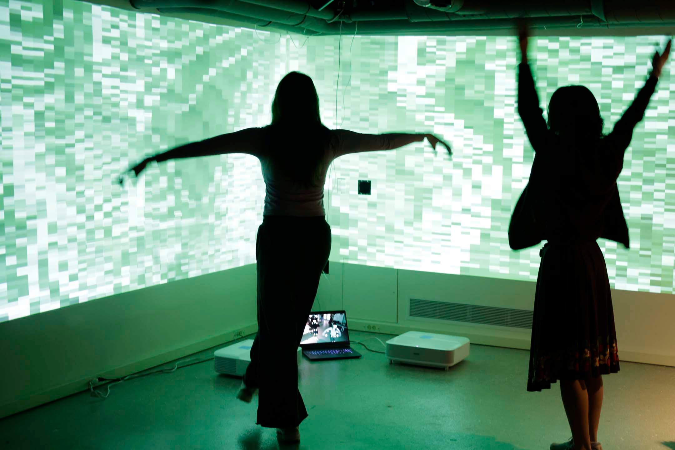 Two people standing in front of a large projection, one is making a T-pose, and the other has their arms in the air. The projection shows a distorted abstract visual that is green and heavily pixelated.